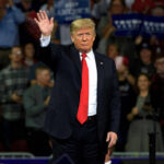 A man in a suit waving to a cheering crowd at a political rally