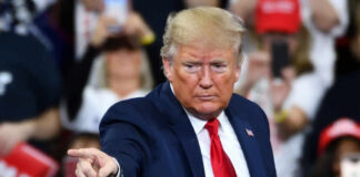 A man in a blue suit and red tie gestures while speaking at a rally