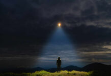 A mysterious figure standing on a mountain under a dark sky with a beam of light from a UFO
