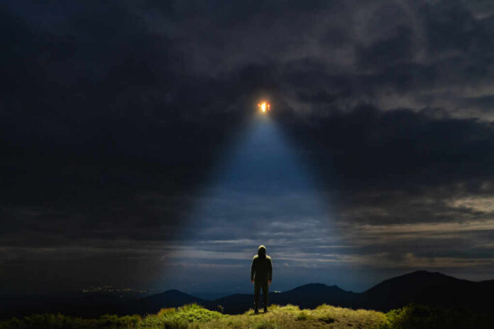 shutterstock_1470669512.jpg A mysterious figure standing on a mountain under a dark sky with a beam of light from a UFO