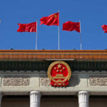 Chinese flags and national emblem displayed on a government building against a blue sky