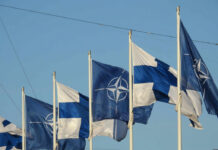 Flags of Finland and NATO waving in the wind against a clear blue sky