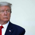 A serious-looking man in formal attire with an American flag pin