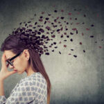 A woman with glasses holding her head, with fragments dispersing from her head