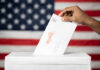 A hand placing a ballot into a voting box with an American flag background