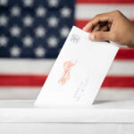 A hand placing a ballot into a voting box with an American flag background