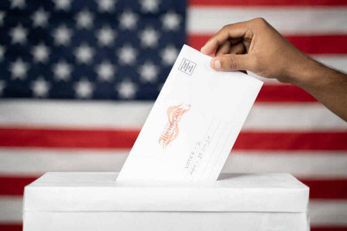 A hand placing a ballot into a voting box with an American flag background