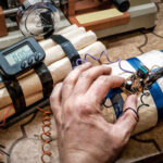 A hand working on a device resembling an explosive with wires and a timer