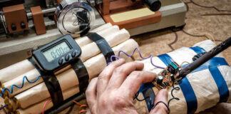 A hand working on a device resembling an explosive with wires and a timer