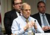 A serious-looking man in a formal shirt and tie sitting at a panel during a government hearing