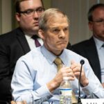 A serious-looking man in a formal shirt and tie sitting at a panel during a government hearing