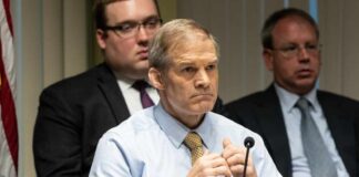 A serious-looking man in a formal shirt and tie sitting at a panel during a government hearing