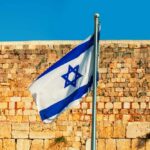 Israeli flag waving in front of a stone wall under a clear blue sky
