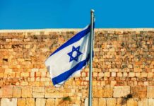 Israel’s Policy Slam—Threatens Holy Land Legacy Israeli flag waving in front of a stone wall under a clear blue sky