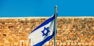 Israeli flag waving in front of a stone wall under a clear blue sky
