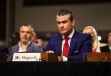 A man in a blue suit and red tie sitting at a hearing table with a nameplate