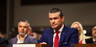 A man in a blue suit and red tie sitting at a hearing table with a nameplate