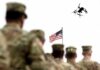 Military personnel standing in formation with a drone flying overhead and an American flag in the background