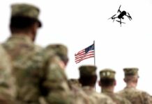 Trump’s Iran Strategy Sparks Constitutional Crisis Military personnel standing in formation with a drone flying overhead and an American flag in the background