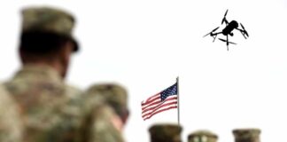 Military personnel standing in formation with a drone flying overhead and an American flag in the background