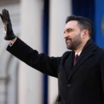 A man in a black coat and gloves waving at an outdoor event