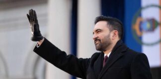A man in a black coat and gloves waving at an outdoor event