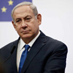 A serious-looking man in a suit at a press conference with a European flag in the background