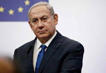 A serious-looking man in a suit at a press conference with a European flag in the background