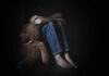 A child sitting on the floor, hugging their knees with a teddy bear beside them in a dimly lit room