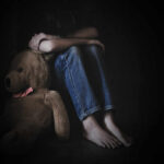 A child sitting on the floor, hugging their knees with a teddy bear beside them in a dimly lit room