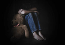 A child sitting on the floor, hugging their knees with a teddy bear beside them in a dimly lit room