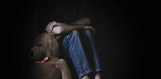 A child sitting on the floor, hugging their knees with a teddy bear beside them in a dimly lit room