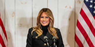 A woman in a black jacket smiling at a podium with a Trump sign and American flags in the background