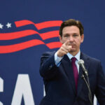 A man in a suit pointing while speaking into a microphone against a backdrop featuring an American flag design