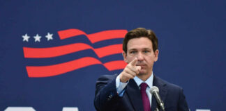 A man in a suit pointing while speaking into a microphone against a backdrop featuring an American flag design