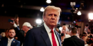 A man in a suit with a red tie stands amidst a crowd of people and cameras