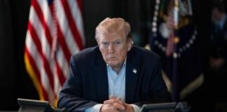 A serious-looking man seated at a table with phones and an American flag in the background