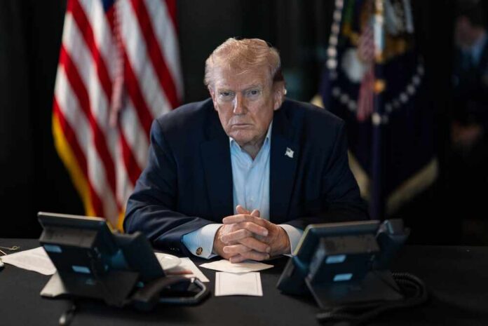 A serious-looking man seated at a table with phones and an American flag in the background