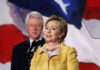 Hillary Clinton speaking at a podium with Bill Clinton in the background