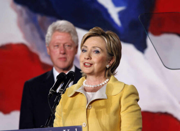 Hillary Clinton speaking at a podium with Bill Clinton in the background
