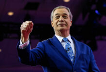 Farage’s £5M Mystery – Under Investigation! A man in a blue suit raising his fist confidently on stage