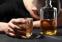 A person with their head down, holding a glass of whiskey next to a bottle