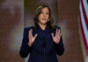 A woman in formal attire delivering a speech at a podium with an American flag in the background