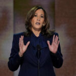 A woman in formal attire delivering a speech at a podium with an American flag in the background