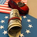 A rolled bundle of cash on a blue background with stars, next to a small red toy car and an American flag.