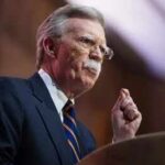 Elite Exemption? Bolton’s Plea Sparks Fury Older man in suit speaking at a podium
