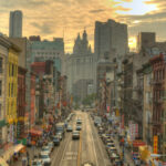 Mayor’s Power Play Freezes Million Rents A bustling street in Chinatown, New York City, with historic buildings and vehicles under a sunset sky