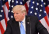 A man in a suit speaking at a podium with American flags in the background