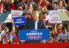 Donald Trump speaking at a political rally with supporters holding signs