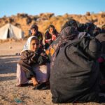 A young girl in traditional clothing sitting among a group of women in a refugee camp